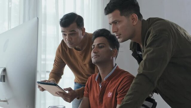 Waist up slowmo of team of male Indian IT specialists looking at computer monitor and having discussion while coworking in office