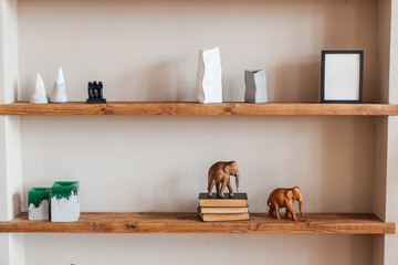 Grey and white colored vases, wooden figures of elephants, blank black photoframe and stack of books on wooden shelf against  grey wall. Home decor.