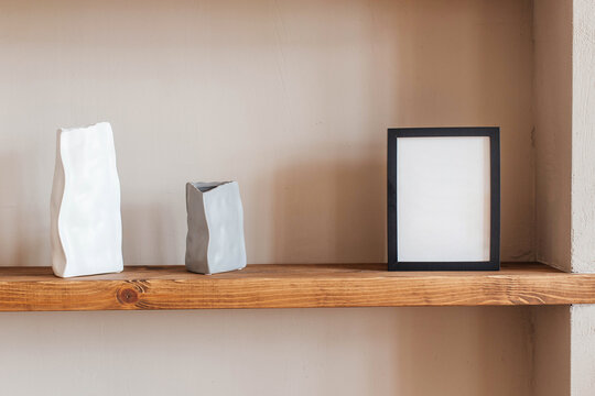Grey and white colored vases, blank black photoframe on wooden shelf against grey wall. Home decor.