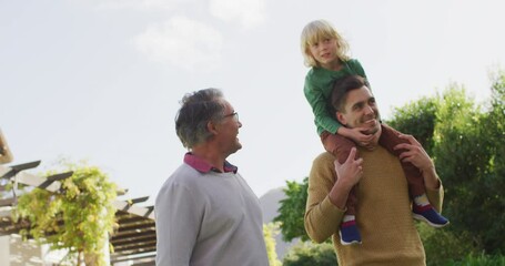 Video of happy caucasian father carrying son on shoulders and walking with grandfather in garden