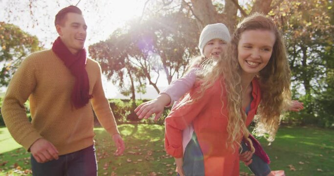 Video of happy caucasian parents, smiling mother piggybacking daughter in sunny autumn garden