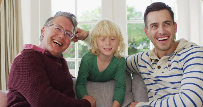 Video portrait of happy caucasian father, grandfather and grandson sitting on couch smiling