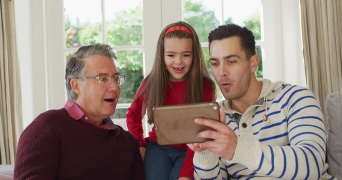 Video of happy caucasian father, grandfather and granddaughter sitting on couch looking at tablet