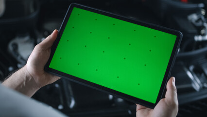 Unrecognizable mechanic holding a tablet with chromakey green screen in his hands, and standing near a car with an opened hood in a car service during a diagnostics