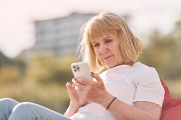 Sitting on the armchair and using smartphone. Senior woman having nice weekend outdoors on the field at sunny day