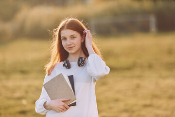 Naklejka premium Student with notepad and with headphones. Young girl is on the field at sunny daytime having nice weekend