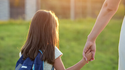 Schoolgirl goes home with mother after school. Girl wearing blue backpack and white dress holds...