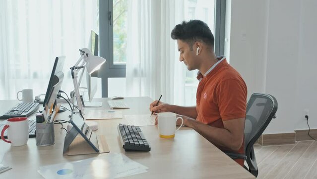 Waist up slowmo of young Indian man in wireless earphones doing paperwork while sitting by computer at office desk