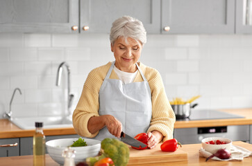 healthy eating, food cooking and culinary concept - happy smiling senior woman with knife chopping red pepper on kitchen at home - Powered by Adobe