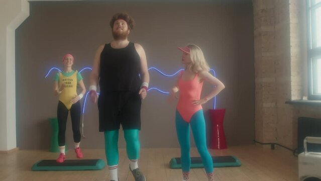 Tracking in of blonde Caucasian female instructor in colorful workout outfit helping young man on group step aerobics class