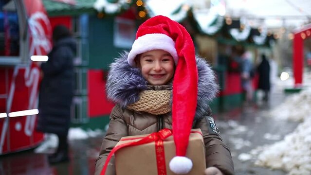 Portrait of joyful girl in Santa hat with gift box for Christmas on city street in winter with snow on festive market with decorations and fairy lights. Warm clothes, knitted scarf and fur. New year