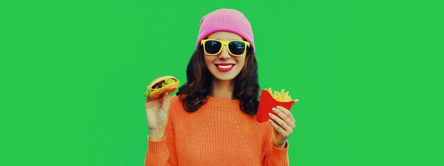 Portrait of stylish happy smiling young woman with burger and french fries fast food wearing pink hat on green colorful background