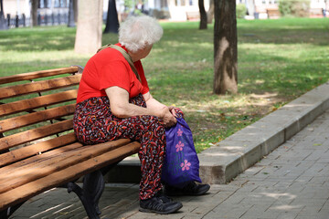 Elderly gray-haired woman sitting with shopping bag on a bench in a park. Leisure in summer city,...