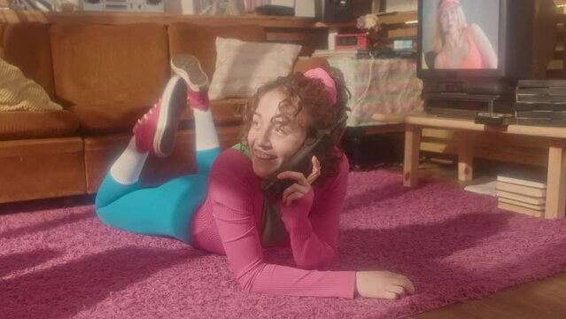 Full shot of young girl in eighties aerobic outfit lying on stomach on floor in living room, talking on phone