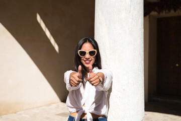 young, beautiful, brunette south american woman is travelling in europe and is leaning on a white marble column while raising the thumbs of her hands giving a thumbs up. Travel and holiday concept.