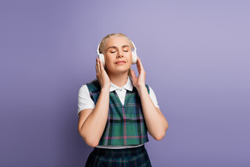 Student in checkered uniform listening music in headphones isolated on purple