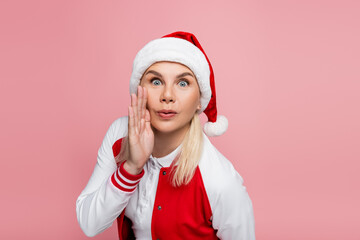 Astonished woman in santa hat holding hand near cheek isolated on pink