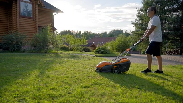 Mature man cuts grass using petrol lawnmower around wooden cottage. Middle-aged person wearing white t-shirt enjoys taking care of private house territory