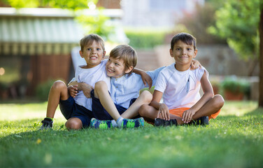 Three happy young boys in summer park. Friends or siblings sitting on grass.