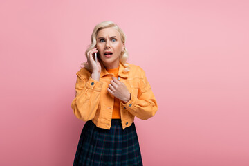 Fototapeta premium Scared blonde woman looking at camera while talking on smartphone isolated on pink