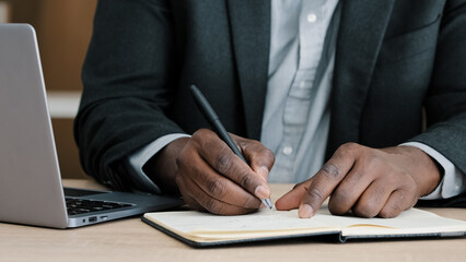Close-up male hands writing in notebook with pen make notes information financial analysis startup project management idea sitting at desk in office using laptop unrecognizable middle-aged businessman