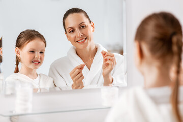 beauty, hygiene, morning and people concept - happy smiling mother and little daughter with dental floss cleaning teeth and looking to mirror at bathroom