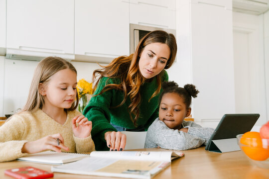 Focused mother helping her daughters with homework on cozy kitchen at home