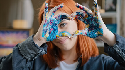 Portrait red-haired girl talented artist female painter stands posing in workshop shows gesture of kindness inspiration love for art sign heart shape with dirty hands in oil acrylic paints smiling