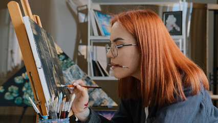Inspired funny focused artist woman with red hair girl painter wears glasses holds painting brushes in mouth paints with oil paints on canvas on easel in studio inspiration creative beautiful drawing