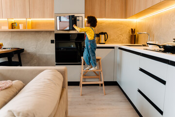Little african boy standing on chair and opening the oven
