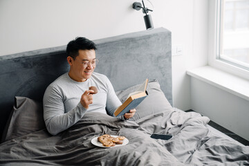 Asian man reading book and eating cookies while sitting in bed