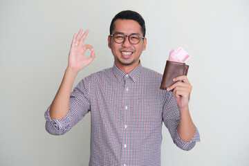 Adult Asian man smiling while holding wallet with money and give OK finger sign