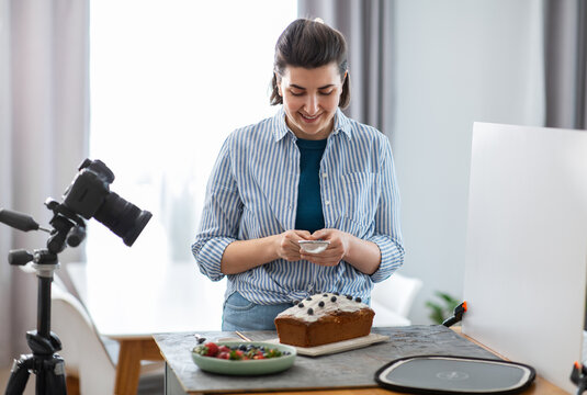 blogging, photographing and people concept - happy smiling female photographer or food blogger with camera pouring powdered sugar to cake in kitchen at home