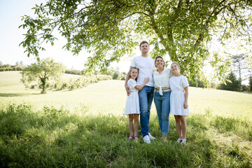 beautiful family with two girls is standing in meadow near walnut tree and has light outfit on and jeans and is happy