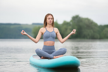 Fototapeta na wymiar Beautiful slender girl doing yoga on a paddleboard. A young woman does water sports in the summer sunrise.