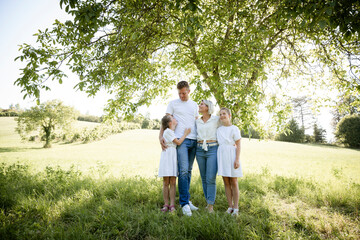 beautiful family with two girls is standing in meadow near walnut tree and has light outfit on and jeans and is happy