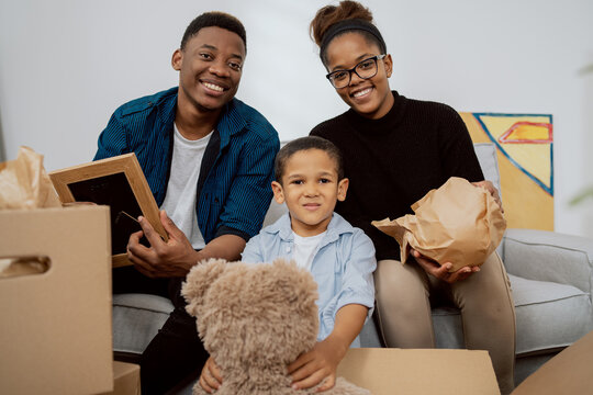 A Loving Family Sits On The Couch In Living Room And Unpacks Boxes After Moving To A New Apartment, They Pull Out Old Memorabilia, Paintings, Parents And Son Smile At The Camera