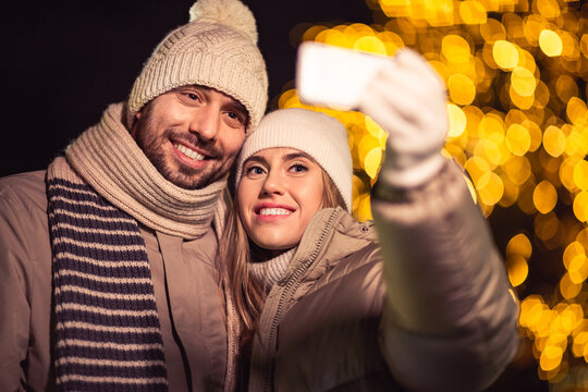 winter holidays and people concept - happy smiling couple taking selfie with smartphone over christmas lights in evening