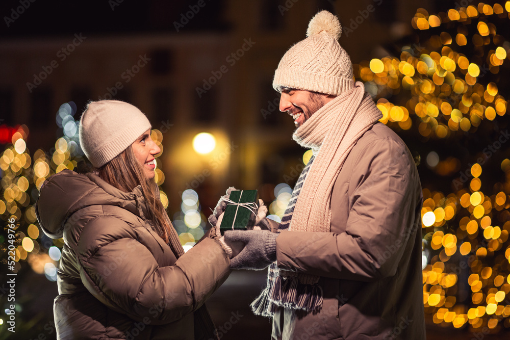 Poster winter holidays and people concept - happy smiling couple with gift over christmas lights in evening