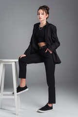 young brunette woman posing with hand in pocket of black trousers near white stool on grey...