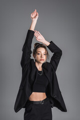 young woman in black crop top and blazer posing with raised hands isolated on grey