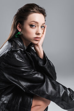 brunette woman in black leather coat touching face and looking at camera isolated on grey
