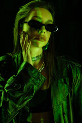 woman in leather coat and fashionable sunglasses touching face in green light isolated on black