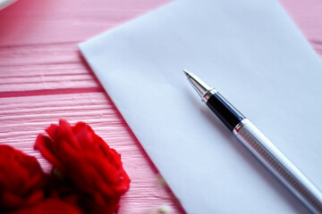 Close up pen on white blank paper and red flowers. Pink wooden background.
