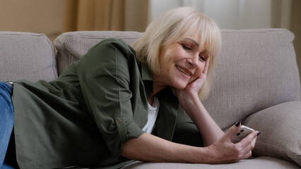 Happy relaxed Caucasian old senior mature woman holding smart phone looking at cellphone laughing lying on sofa couch enjoying using mobile apps for shopping having fun watching funny video in media