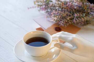 White mannequin hand with cup of coffee. Envelope and flowers.