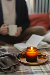 Burning candle on background of young woman holding mug coffee or tea, reading book while sitting in lotus pose on bed in cozy bedroom