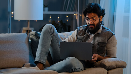 Businessman guy Arab Indian man bearded male user wearing glasses sitting on sofa at night late time with laptop reading bad news online trouble app job problems pondering solution looking at screen - obrazy, fototapety, plakaty