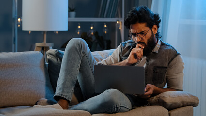 Businessman guy Arab Indian man bearded user wearing glasses sitting on sofa at night late time with laptop reading difficulties news online trouble thinking idea app pondering solution looking screen
