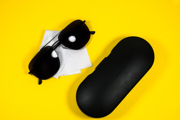 Black frame mens sunglasses with glasses cleaning cloth and case on a yellow background.